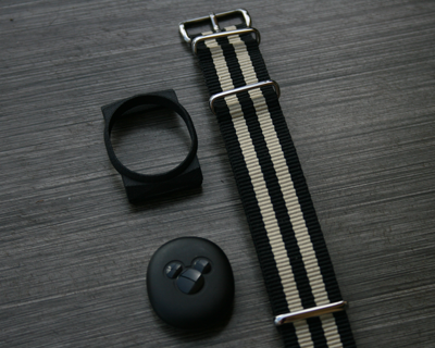 A nylon band sits on a grey table, next to a 3D-Printed harness for a plastic puck, which sits below.